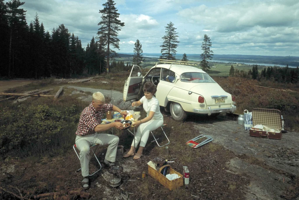 Ett par äter lunch på en utkiksplats på 60-talet. Bilen står parkerad på en klipphäll och det är fin utsikt över en sjö.