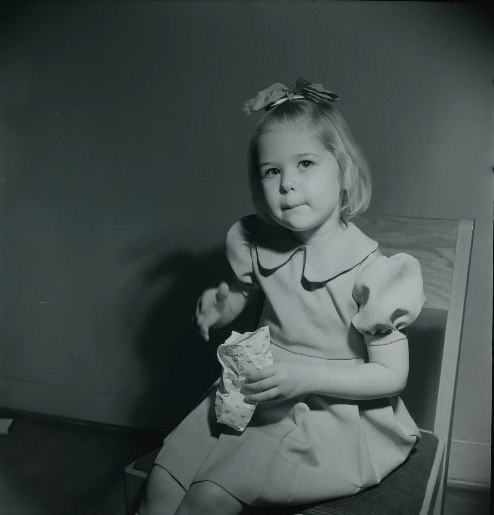 En flicka i klänning och rosett i håret sitter på en stol och äter godis, hon håller en godispåse i handen.