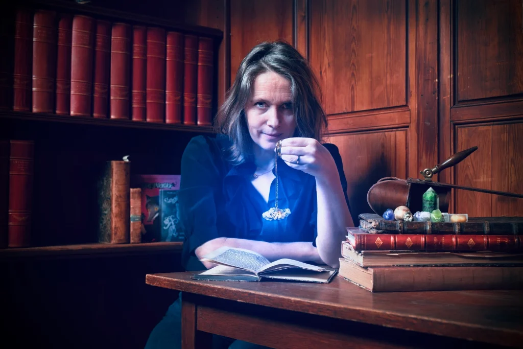 En kvinna sitter vid ett bord med en bokhylla med böcker bakom sig. Hon håller i en magisk pendel med ljuseffekt i blått och har en bok framför sig.