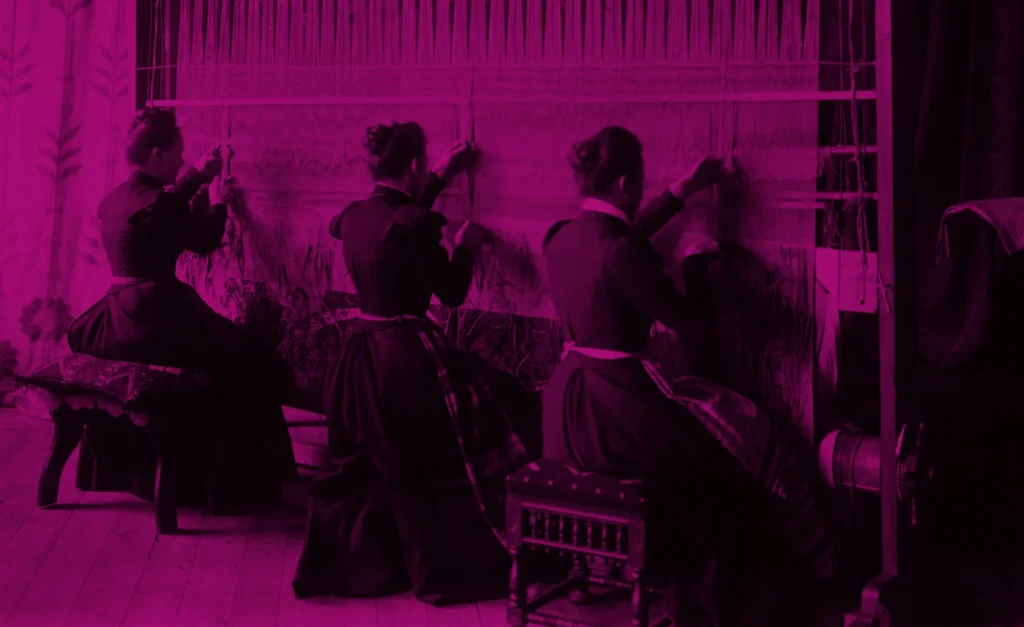 Tre kvinnor i klänningar och uppsatt hår som sitter ner på pallar och arbetar på en textilfabrik.