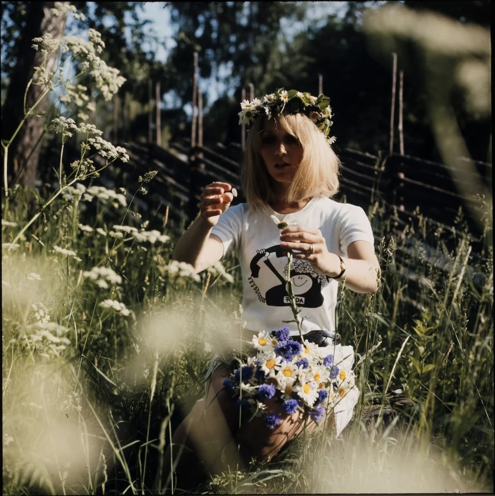 En blond kvinna i t-shirt med krans i håret plockar blommor. En gärdsgård i bakgrunden.