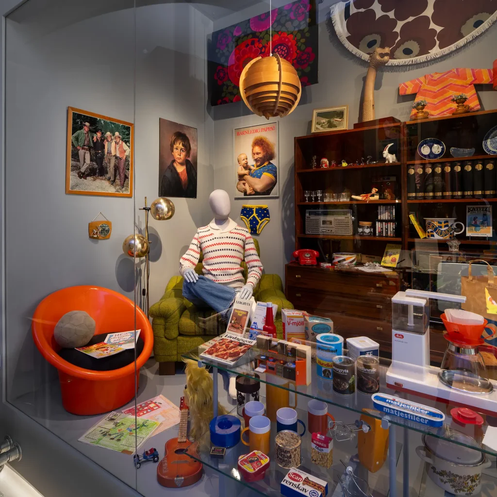 En monter som liknar ett vardagsrum från 1970-talet med bokhylla, orange plastfåtölj, bilder på väggarna och mängder av vardagsföremål som köks- och hushållsprylar, matförpackningar och LP-skivor.