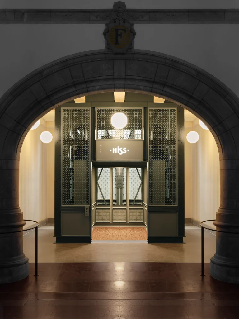 Genom en valvportal syns en hiss med dekorerad järnsmide där Nordiska museets symbol bilder ett mönster, hissdörrarna står öppna.