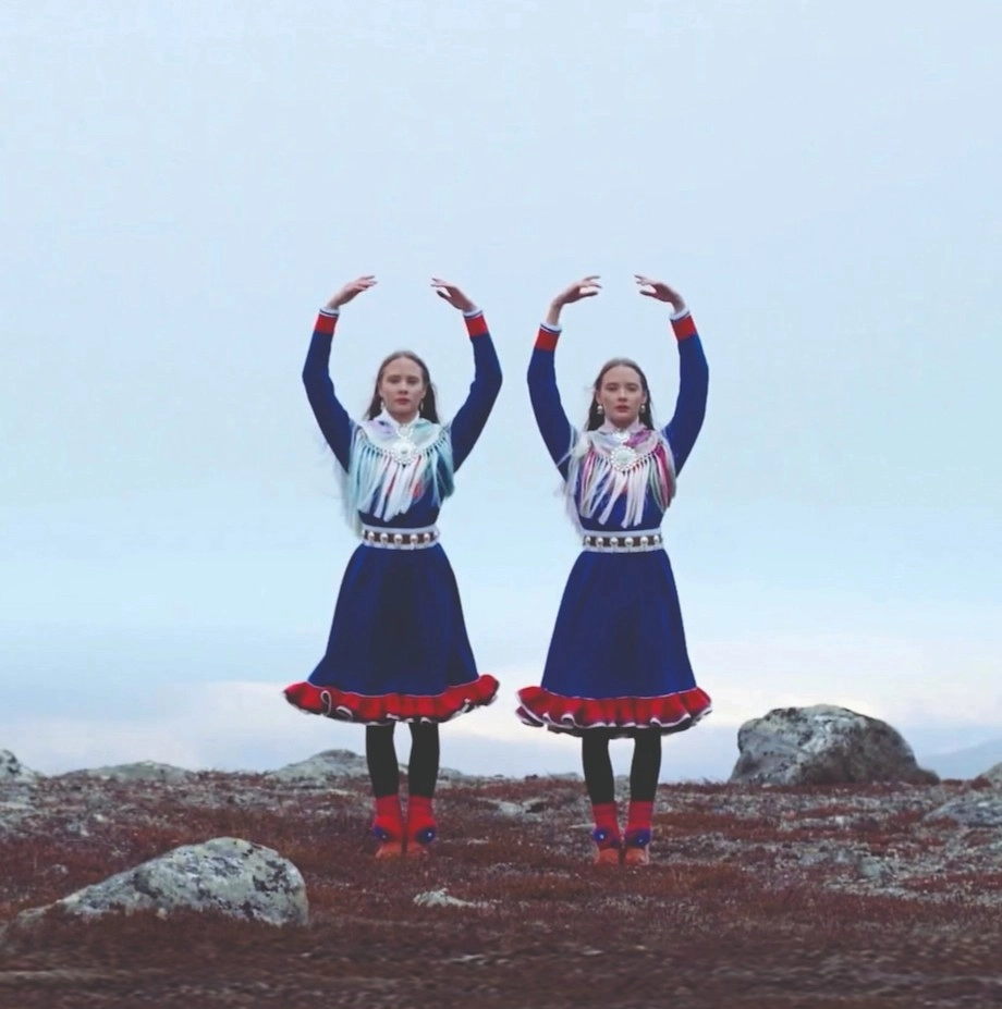 Två unga dansöser i samiskinspirerade dräkt står på en karg hed mot ljus himmel bakom sig och sträcker armarna i en en haut.