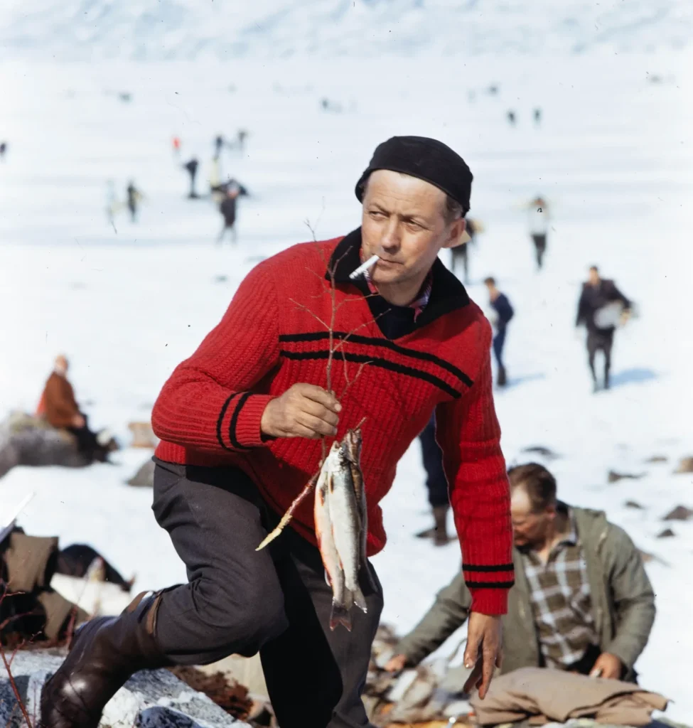 En man pimpelfiskar utanför Torneträsk runt 1970. Han har precis fått upp en fisk.