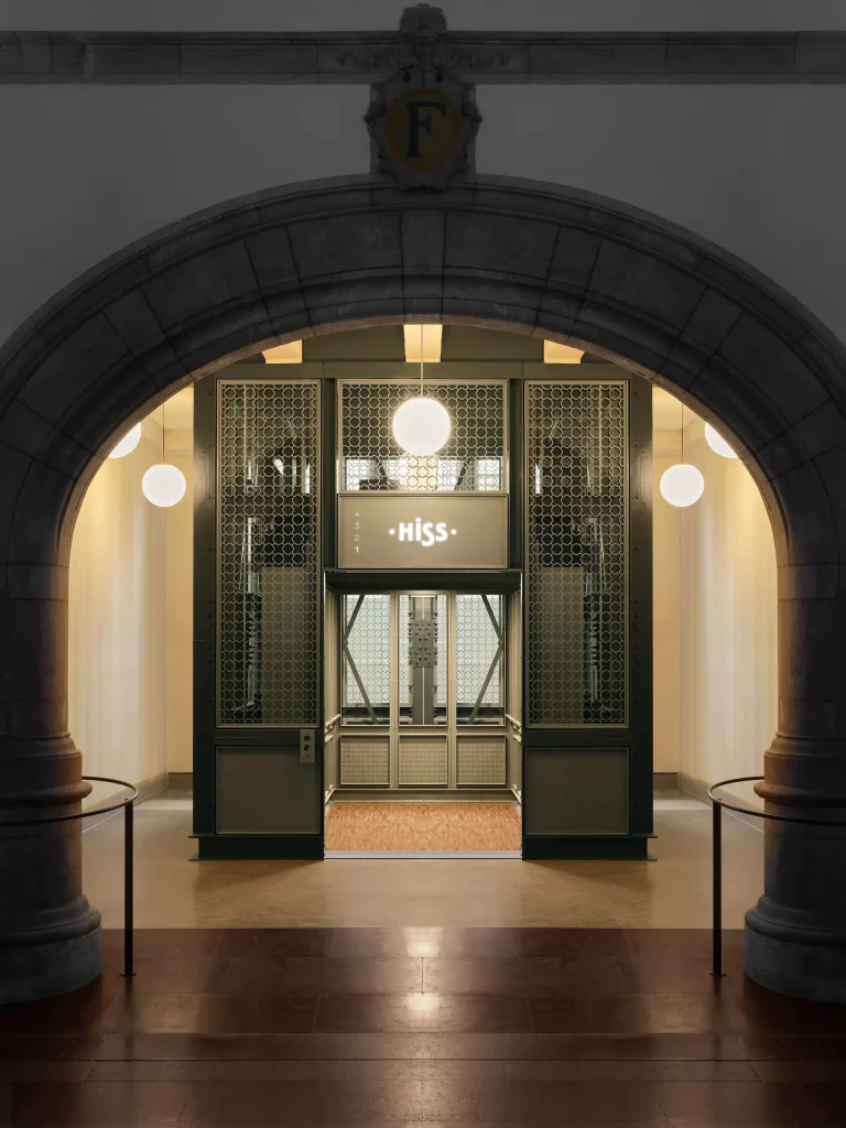 En bild av en hiss med öppen dörr, den har en olivgrön ton och järnsmidesutsmyckning i form av ett mönster bildat av Nordiska museets symbol.