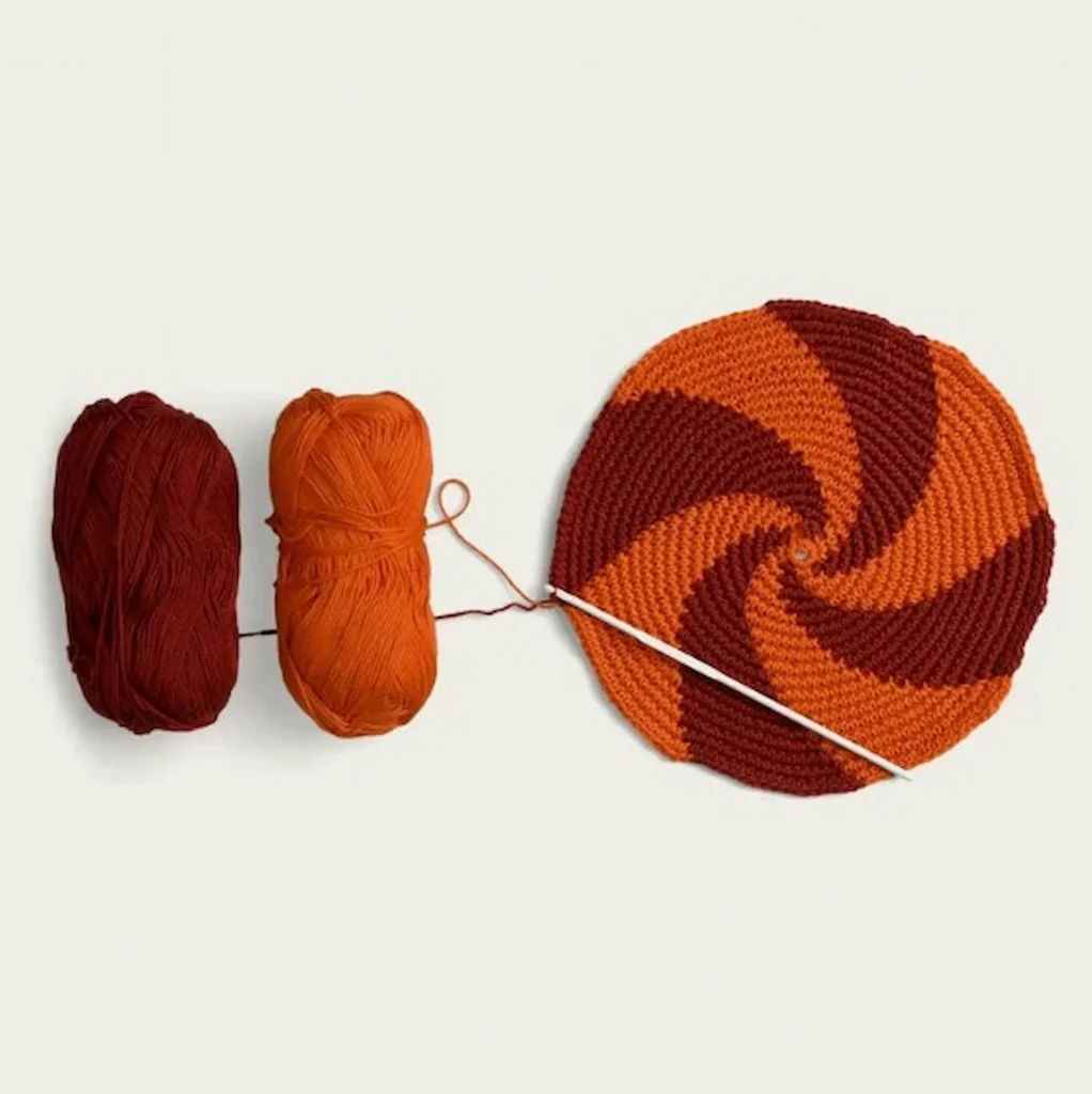 Två nystan och en virkad grytlapp i orange och vinrött mot vit bakgrund