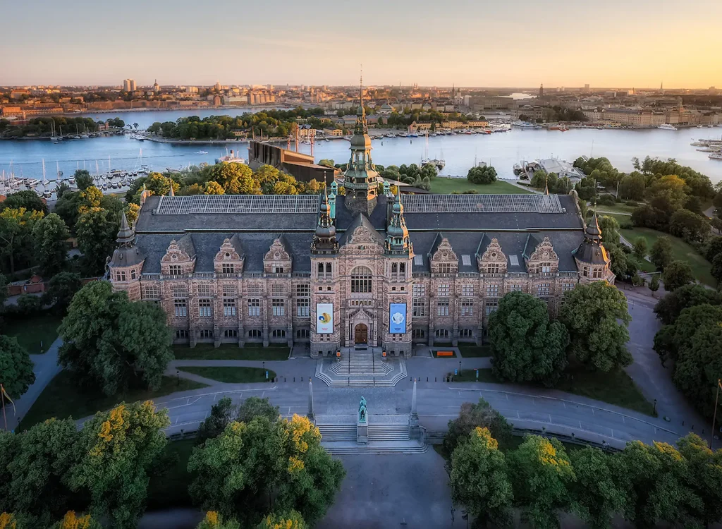 En palatslik byggnad med tinnar och torn sedd uppfrån med vatten, grönska och Stockholm runtom