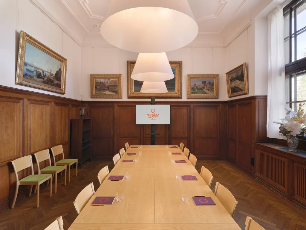 Ett mötesrum med mörk träpanel, parkettgolv, stort fönster, konferensbord och storbildsskärm med Nordiska museets logotyp