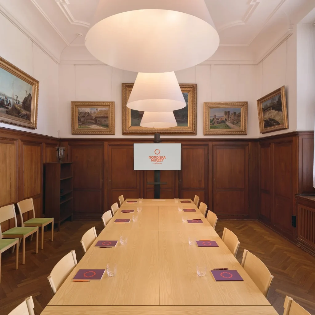 Ett mötesrum med mörk träpanel, parkettgolv, stort fönster, konferensbord och storbildsskärm med Nordiska museets logotyp