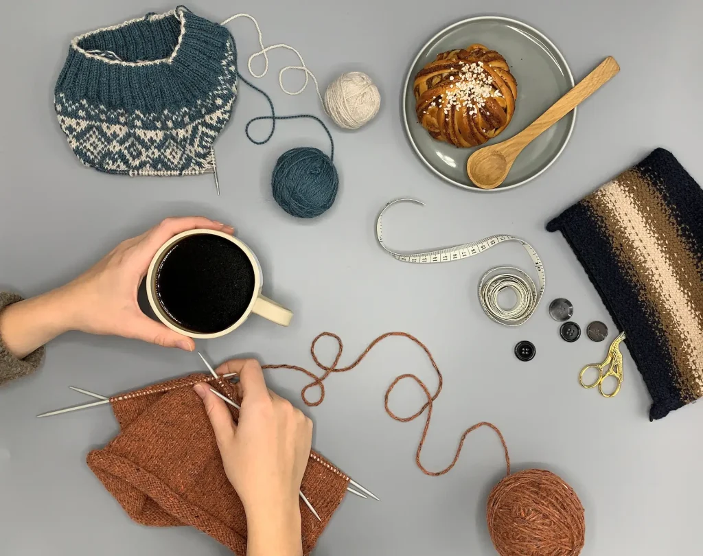 En hand håller i en kopp kaffe och en stickning, bredvid syns en kanelbulle och handarbeten
