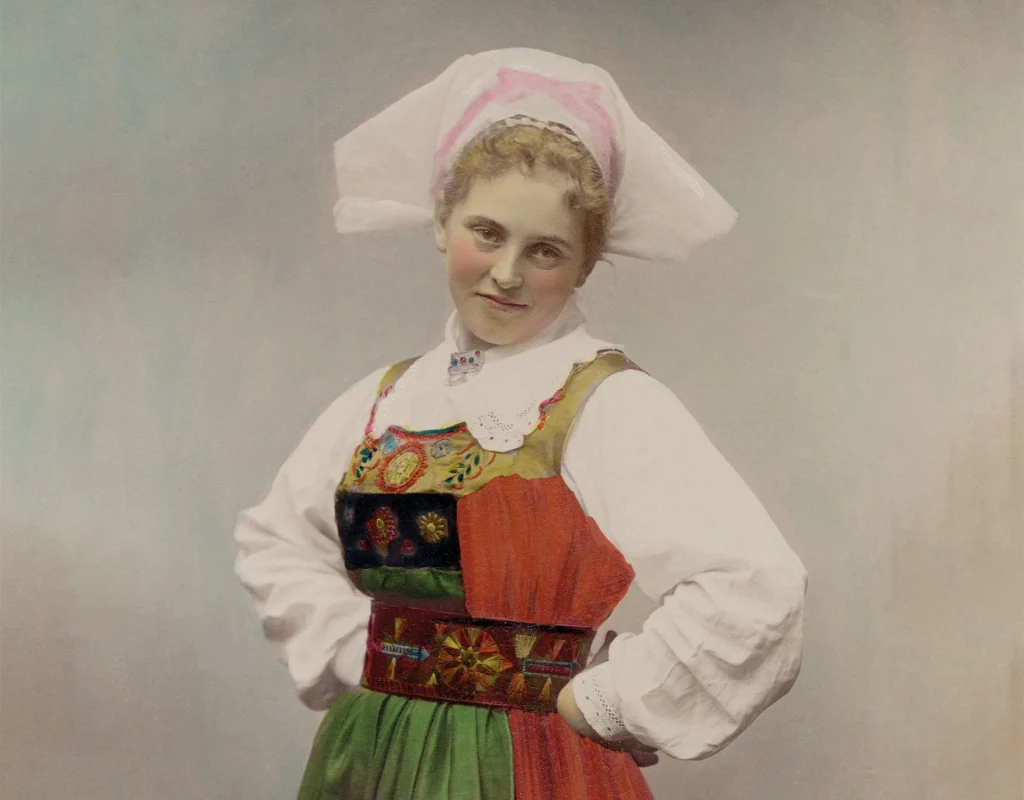 Ett kolorerat fotografi av en kvinna i folkdräkt med händerna kaxigt i sidorna.