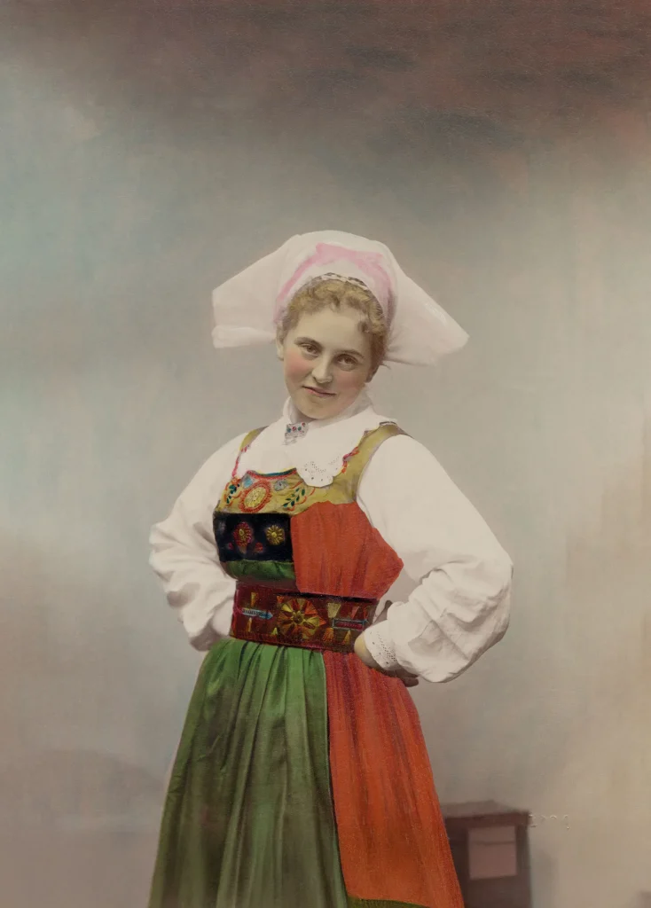 Kolorerat äldre fotografi av en ung kvinna i folkdräkt, hon står kaxigt med händerna i sidorna och ler mot kameran.