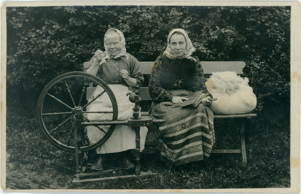 Ett äldre fotografi på två kvinnor som sitter på en bänk med sin spinnrock och ull.