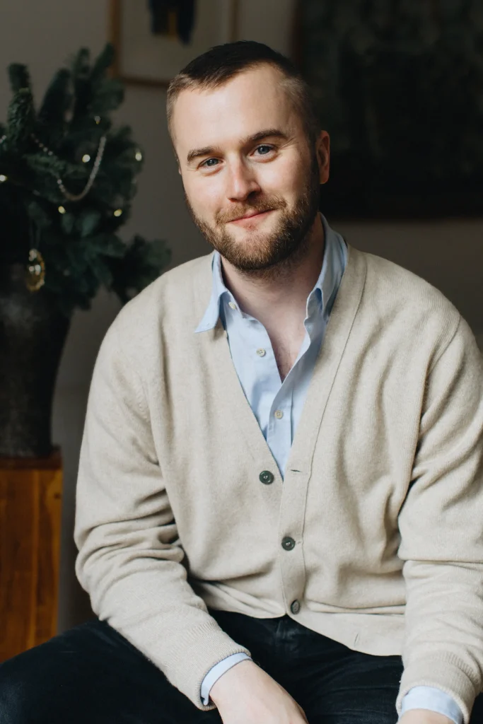 En ung man med skägg, blå skorta och beige cardigan ler mot kameran, bakom honom skymtar en bordsgran.
