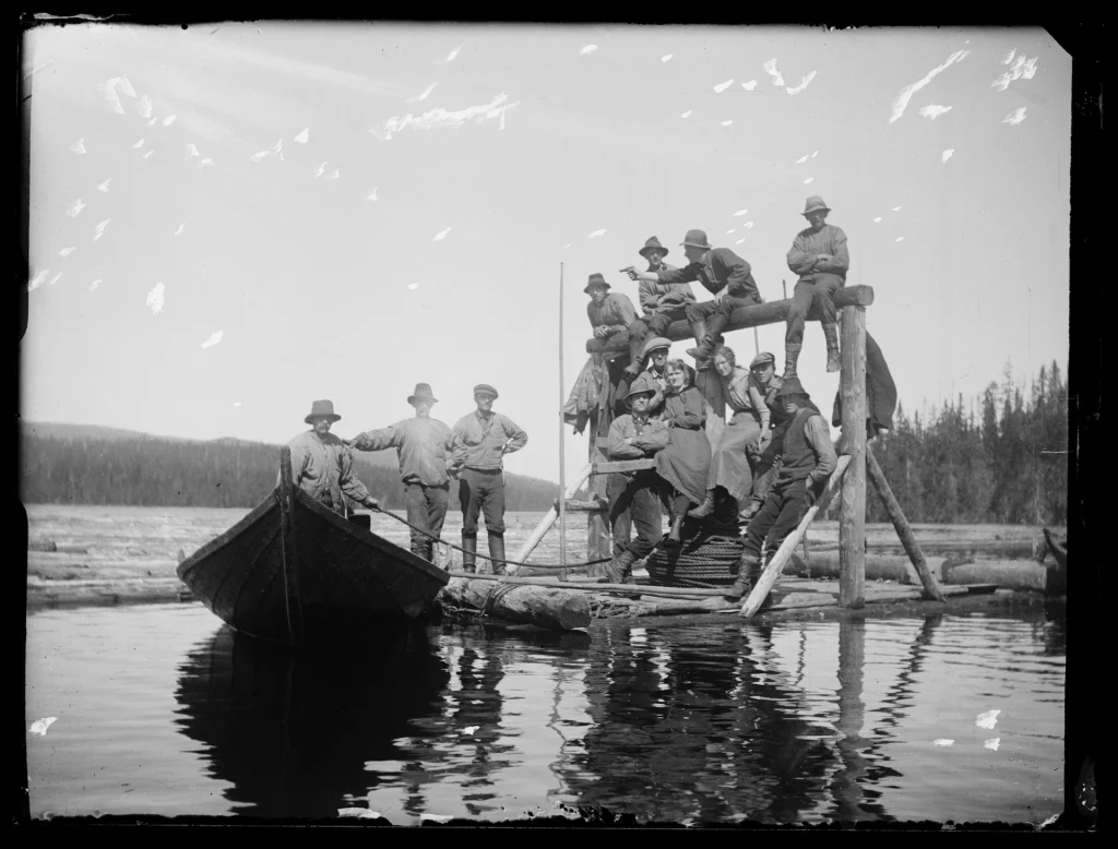 Flera timmerflottare sitter på en brygga vid Almsjön i Dalarna. Vid bryggan är en liten båt förtöjd.