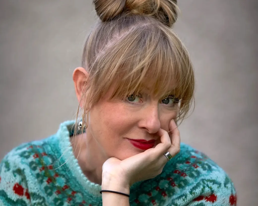 En kvinna med håret i knut och med mönstrad stickad tröja ler mot kameran.