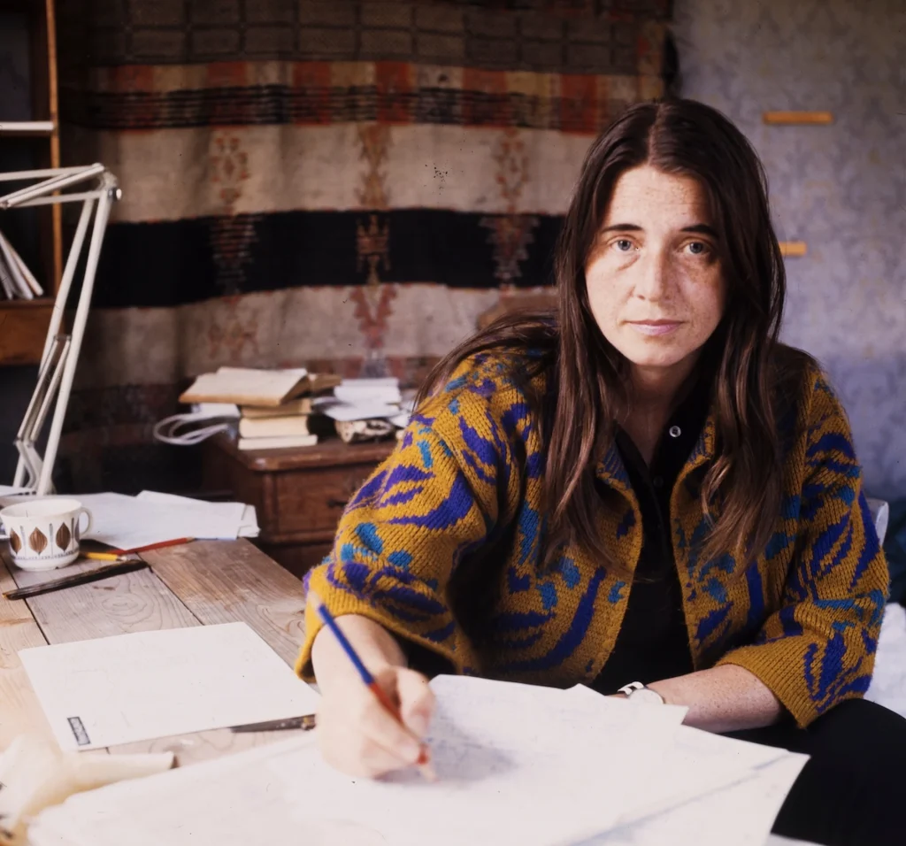 En kvinna med långt mörkt hår och mönstrad kofta sitter vid ett skrivbord och skriver på ett papper