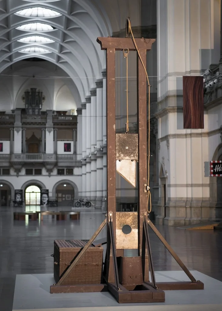 I en stor hall står en hög giljotin. I en lång lina hänger bilan som är diagonalt i änden, en rund öppning förhuvudet syns och under den ett zinkkar för att ta emot huvudet.