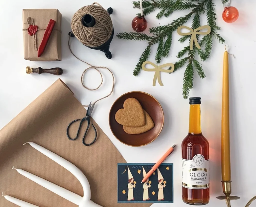 Julpapper, lackstång, glöggflaska, pepparkakor, stearinljus ligger i ett stilleben på en vit yta