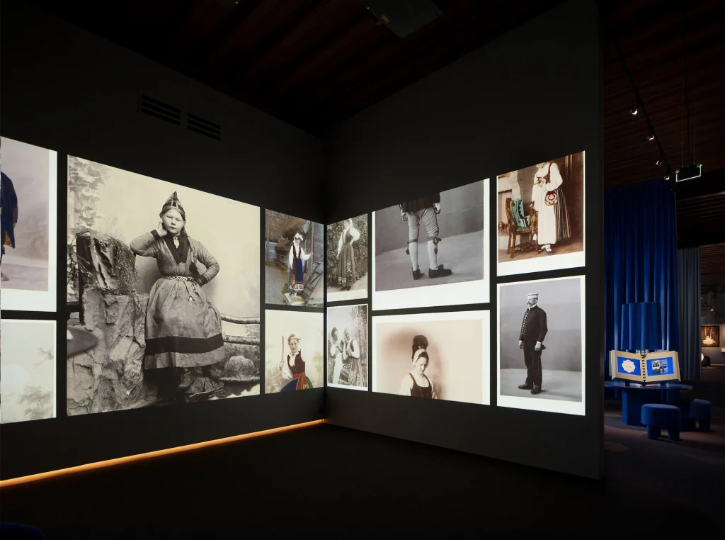 Ett mörk rum med bilder i storformat projicerade i ett bildflöde längs väggarna, till höger syns fortsättningen av utställningen som är i flera rum i fil.