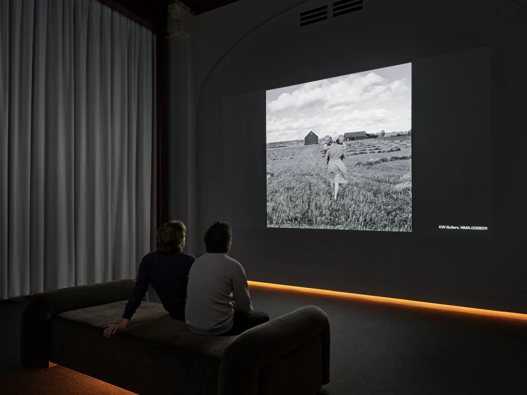 Två män sitter på en divan och tittar på ett fotografi av en kvinna och två barn som projiceras på vägg.