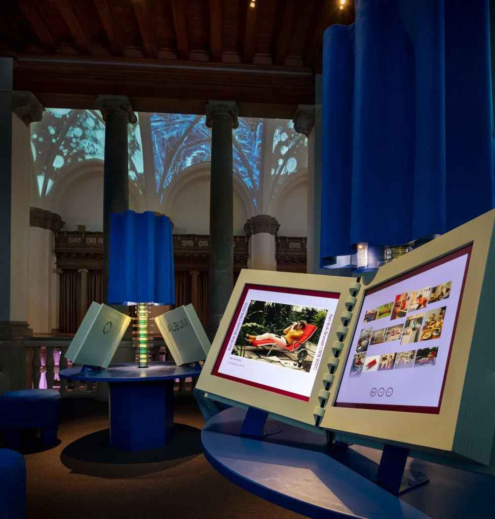 Ett rum med kolonner, blå draperier och runda bord med digitala fotoalbum att klicka och bläddra i, i bakgrunden utsikt mot en stor hall med kryssvalv med takprojektioner.