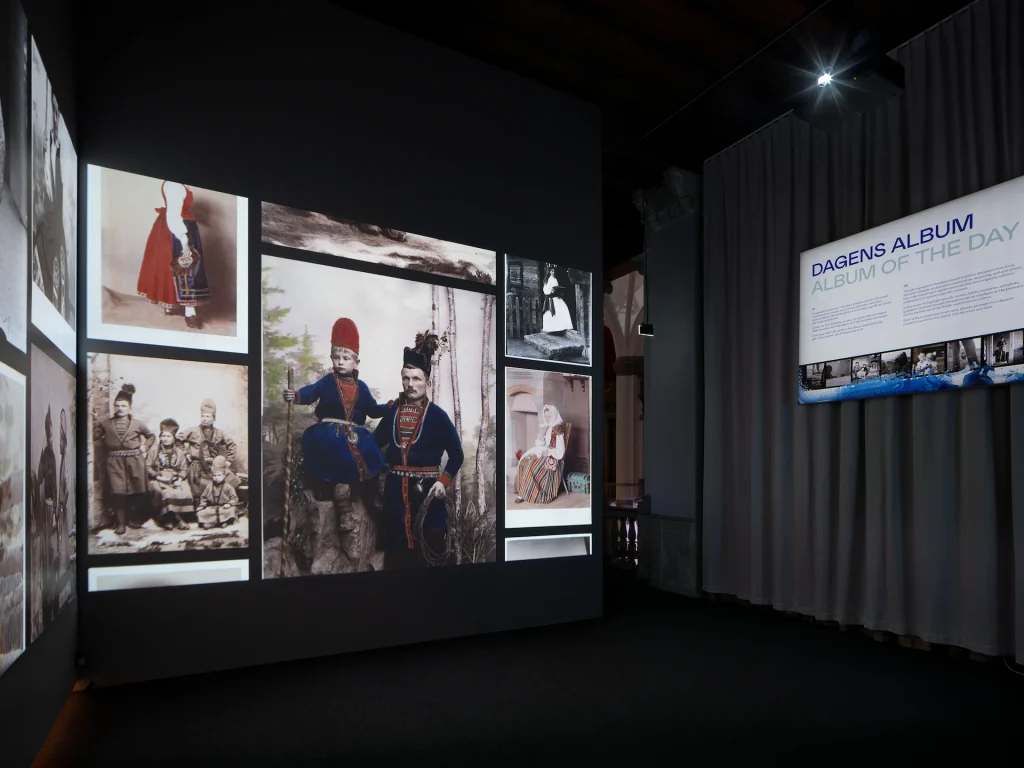 Ett mörk rum med bilder i storformat projicerade i ett bildflöde längs väggarna, en digital skylt med texten DAGENS ALBUM.
