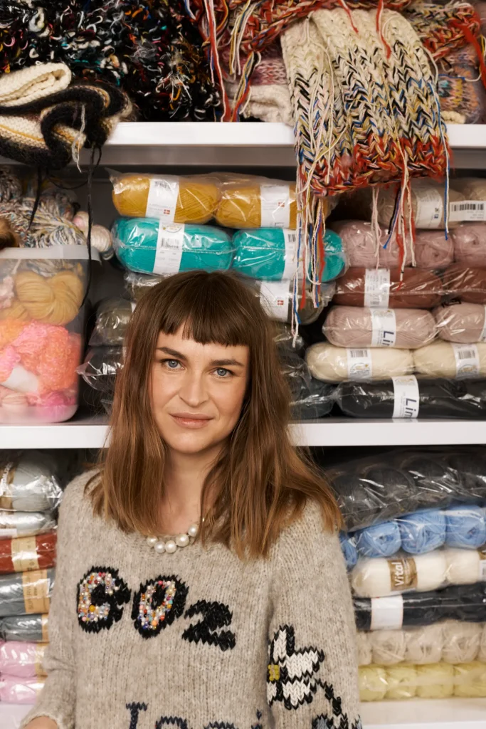 En kvinna med stickad tröja står vid en bokhylla full med garnnystan i starka färger