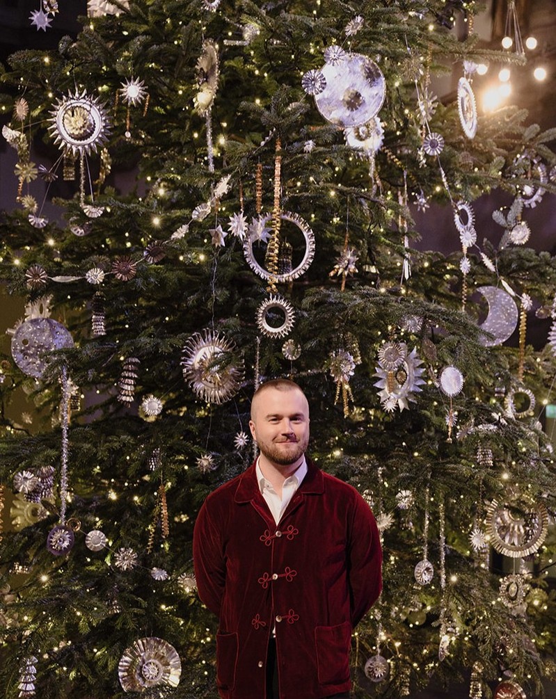 En man i vinröd sammetsjacka står framför en enorm gran klädd i dekorationer av kakformar, stjärnor, stjärntecken, solar och stjärnfall.