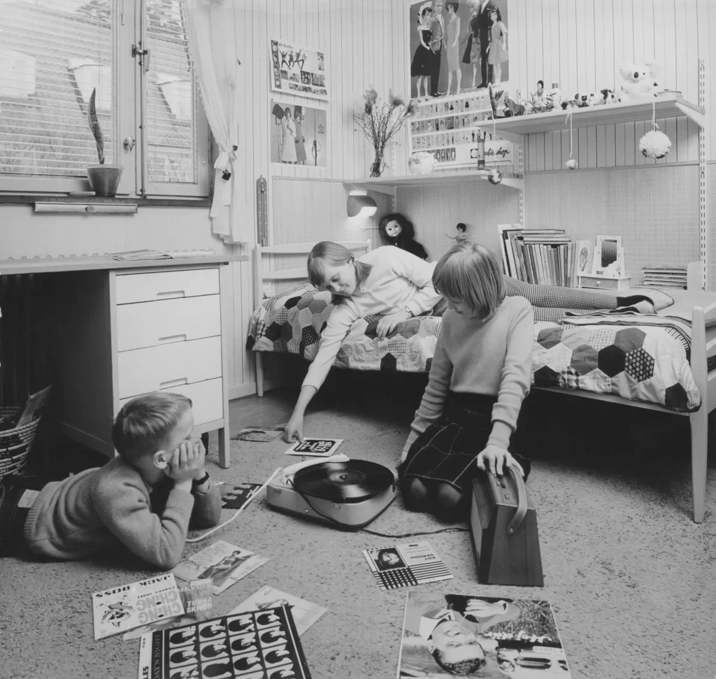 Tre barn i olika åldrar i ett barnrum med säng, skrivbord, leksaker och grammofon.