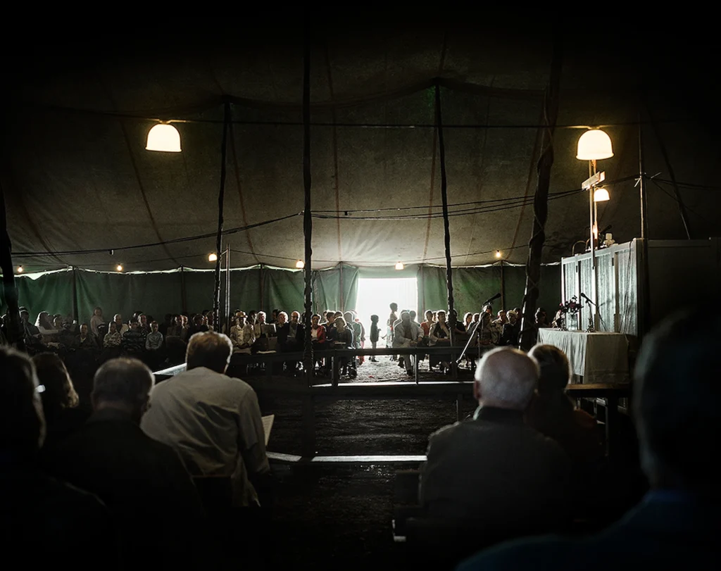 Tältmöte med en församling, det är mörkt i tältet trots lampor i taket, till höger ett altare med träkors