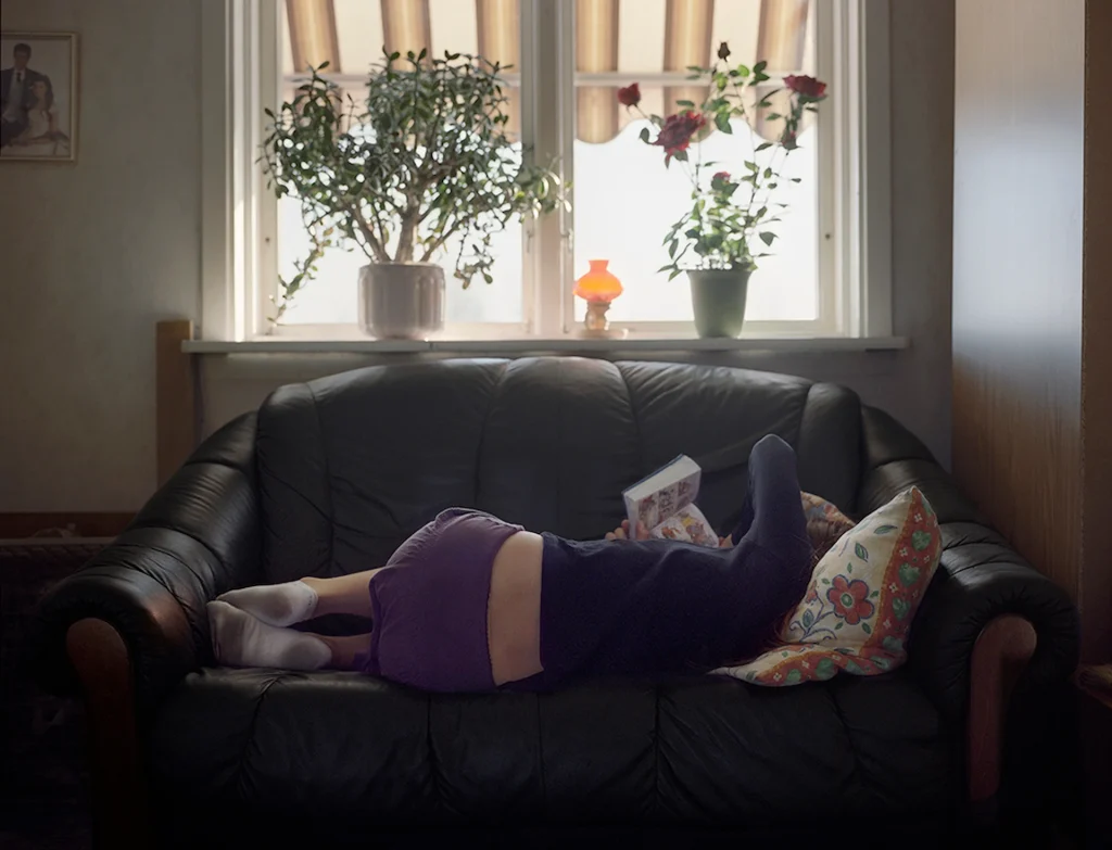 En ung tjej ligger i en soffa vid ett fönster och läser en seriepocket med ryggen vänd från rummet för att få en stund ifred