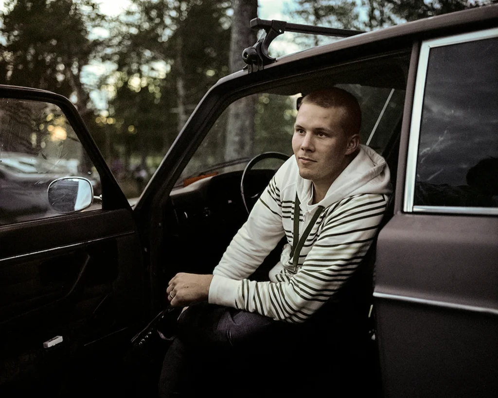 En ung kille sitter i framsätet vänd från ratten i en bil med öppen dörr, i bakgrunden skog och kvällsol