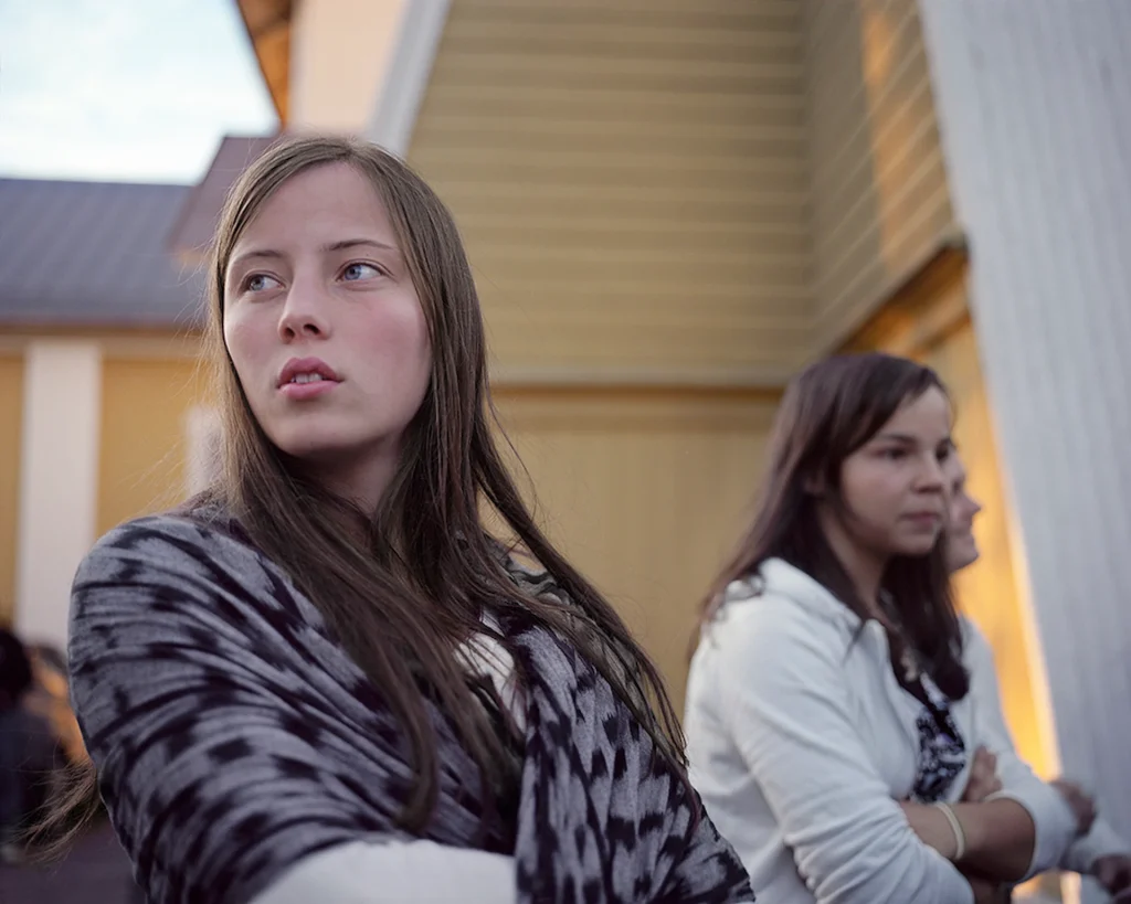 En ung tjej med långt hår och en mönstra sjal om axlarna blickar bort i fjärran, i bakgrunden, fler ungdomar