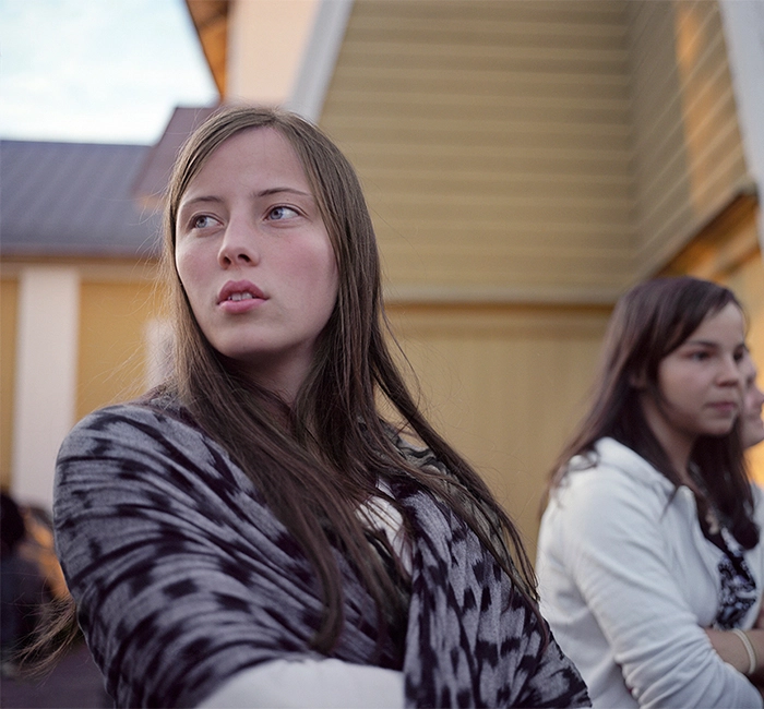 En ung tjej med långt hår och en mönstra sjal om axlarna blickar bort i fjärran, i bakgrunden, fler ungdomar