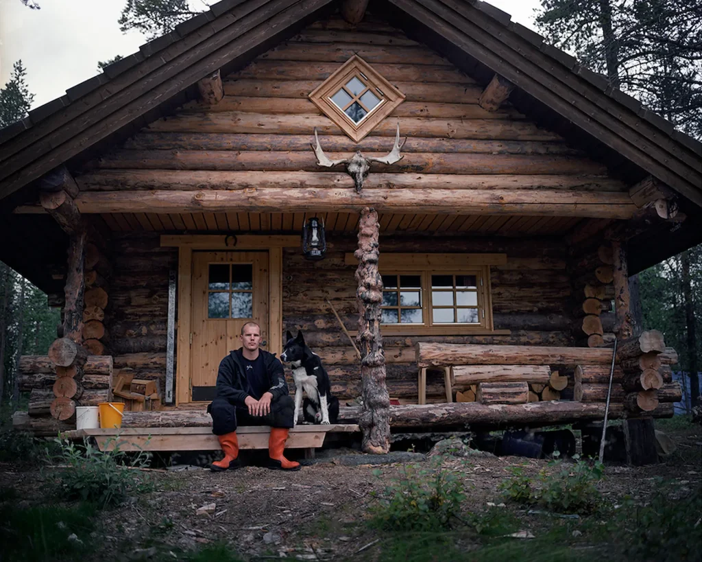 En man i röda gummistövlar sitter på bron till en timmerstuga, bredvid sitter honom en stor svart och vit hund