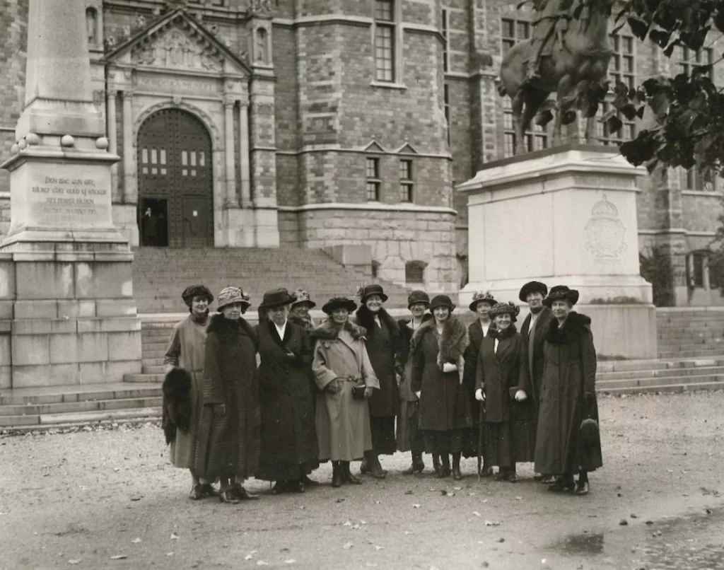 En grupp om tolv damer i långa kappor och hattar står uppradade och leende nedanför trappan till Nordiska museet år 1923.