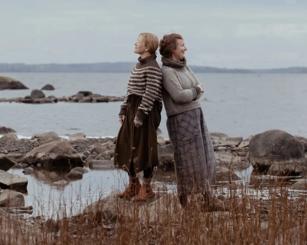 Två kvinnor står rygg mot rygg i strandkanten iklädda stickade tröjor och vida kjolar.