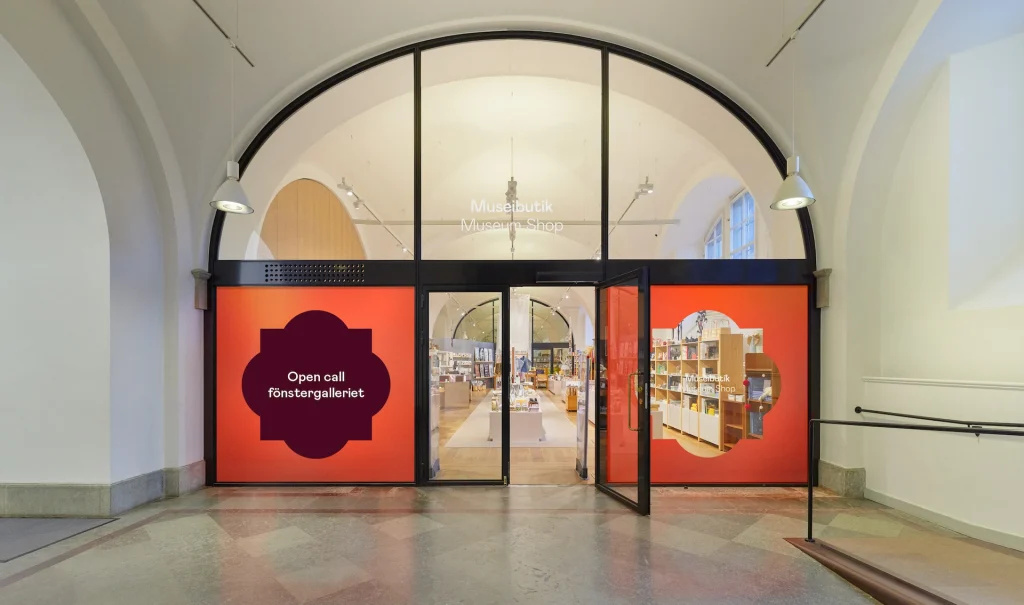 En ljus entréhall med höga valv och glasad ingång till en museibutik. Glasväggarna har en orange inramning i form av Nordiska museets symbol. Ena fönstret är ifyllt med texten open call - museibutikens fönstergalleri.