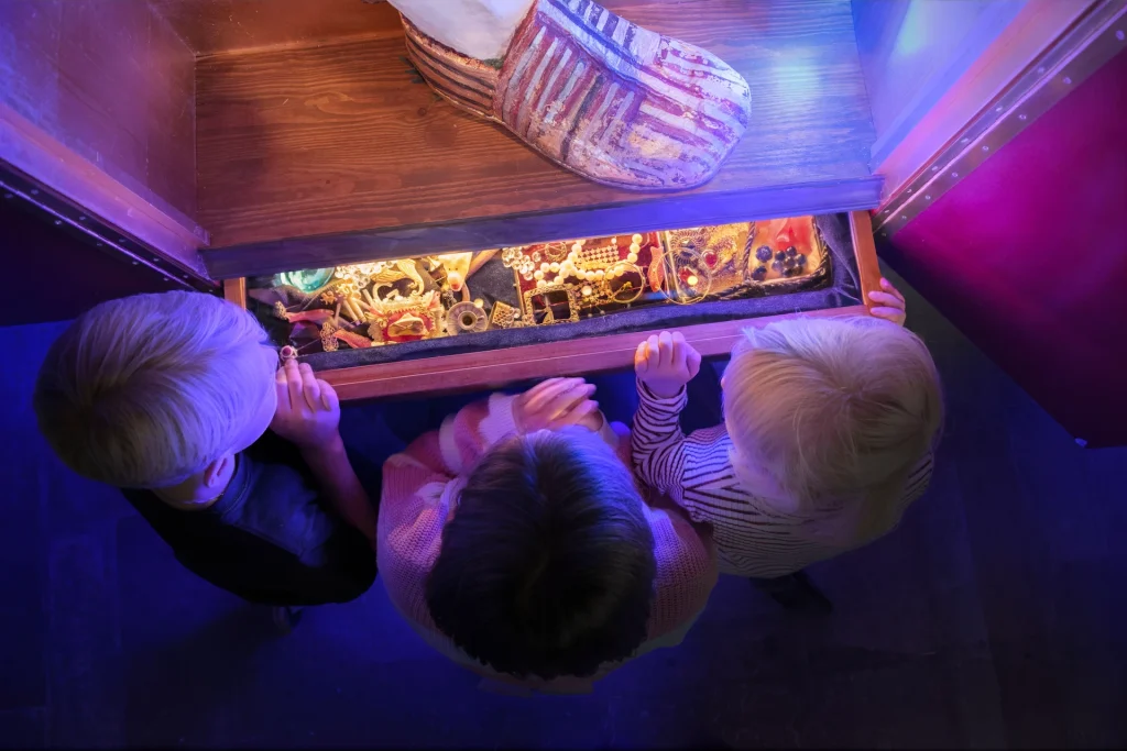 Tre barn uppifrån som kikar ner i en låda fylld med glittrande skatter